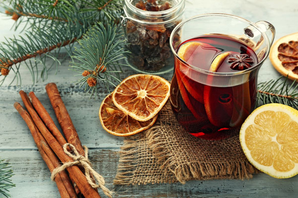 KI generiert: Das Bild zeigt eine weihnachtlich-winterliche Szene mit einem Glas Glühwein, das von getrockneten Orangenscheiben, Zimtstangen, Tannenzweigen und Zitronenscheiben umgeben ist. Im Hintergrund befindet sich ein Glas mit Rosinen auf einem rustikalen Holztisch.