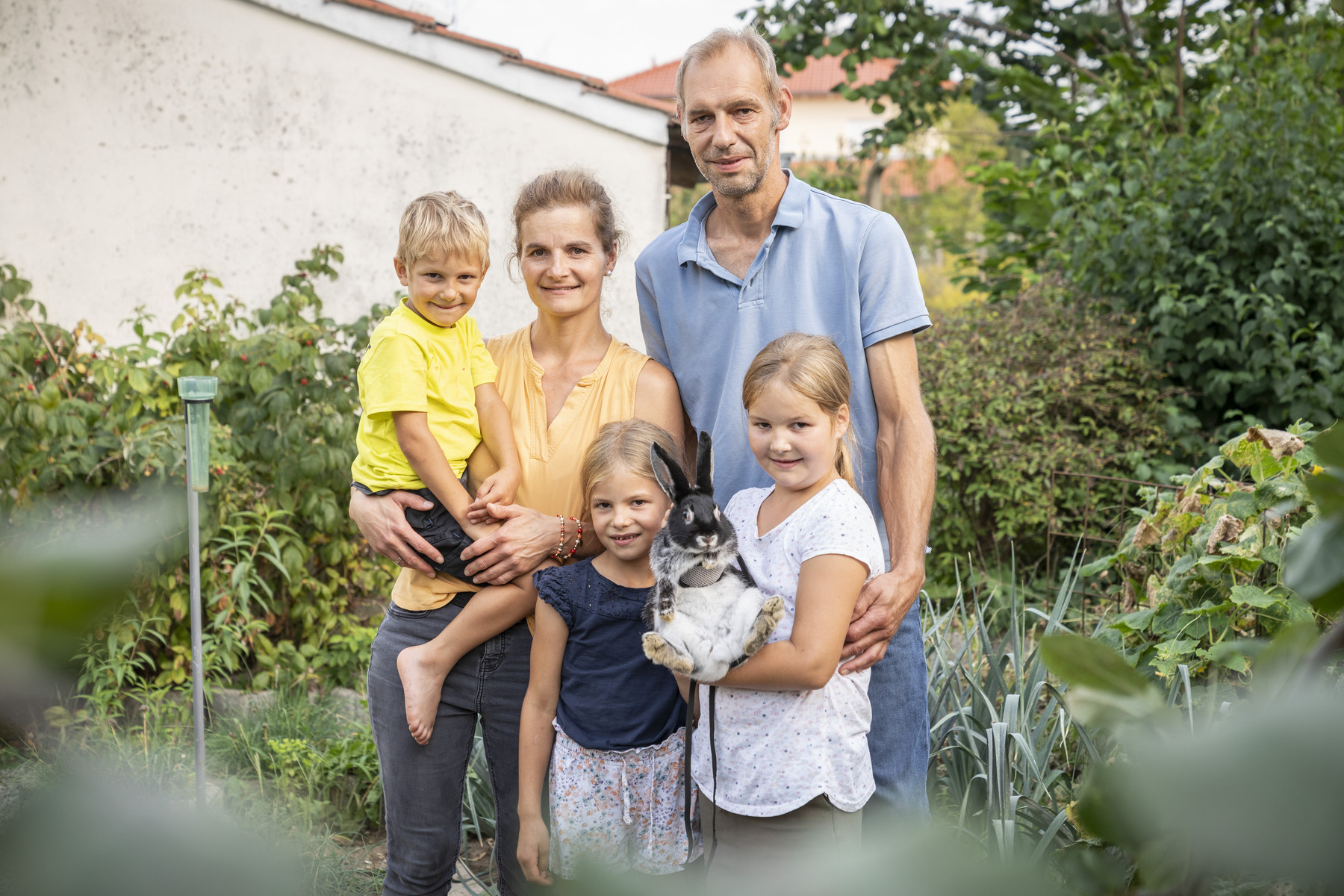 KI generiert: Eine Familie steht in einem Garten und lächelt in die Kamera, während eines der Kinder ein Kaninchen hält. Im Hintergrund sind Pflanzen und ein Gebäude zu sehen.