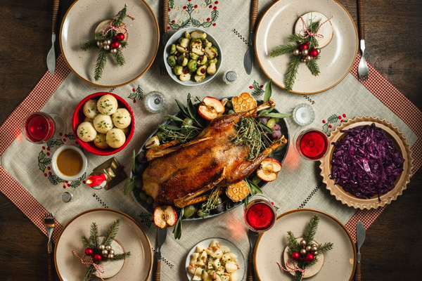 KI generiert: Das Bild zeigt einen festlich gedeckten Tisch mit einem gebratenen Gänsebraten als Hauptgericht, umgeben von verschiedenen Beilagen wie Rotkohl, Kartoffelknödeln und Rosenkohl. Der Tisch ist dekoriert mit saisonalen Elementen, einschließlich Kerzen und Tannenzweigen.