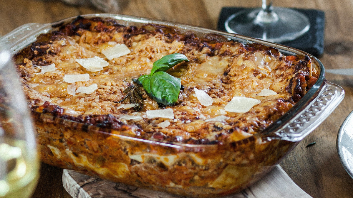 KI generiert: Das Bild zeigt eine frisch gebackene Lasagne in einer Glasauflaufform, garniert mit frischem Basilikum und Parmesansplittern. Im Hintergrund sind ein Weinglas und eine bessere Tischdekoration erkennbar.