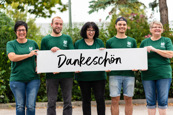 KI generiert: Eine Gruppe von fünf Personen steht draußen und hält ein Schild mit der Aufschrift "Dankeschön". Alle tragen grüne T-Shirts und lächeln in die Kamera.