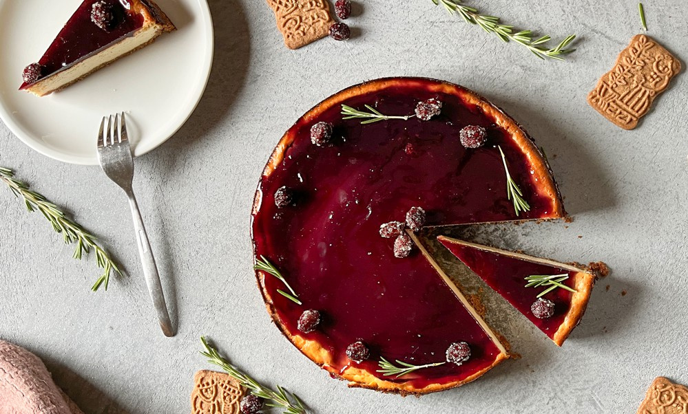 KI generiert: Das Bild zeigt einen Kuchen mit einer glänzenden, roten Glasur und dekoriert mit Rosmarinzweigen und Beeren. Ein Stück des Kuchens wurde herausgenommen und liegt auf einem Teller neben dekorativen Keksen.