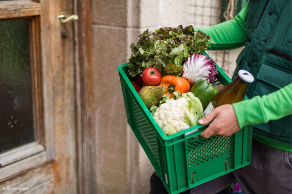 KI generiert: Das Bild zeigt eine Sortierhalle, in der Arbeiter Gemüse in grüne Kisten verpacken. Im Hintergrund ist ein Transparent mit dem Logo "Die rollende Gemüsekiste" zu sehen.