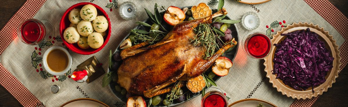 KI generiert: Das Bild zeigt einen festlich gedeckten Tisch mit einem gebratenen Gänsebraten als Hauptgericht, umgeben von verschiedenen Beilagen wie Rotkohl, Kartoffelknödeln und Rosenkohl. Der Tisch ist dekoriert mit saisonalen Elementen, einschließlich Kerzen und Tannenzweigen.